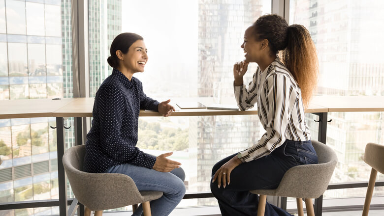 Two professional women sit facing each other in an office with large windows, engaged in conversation.